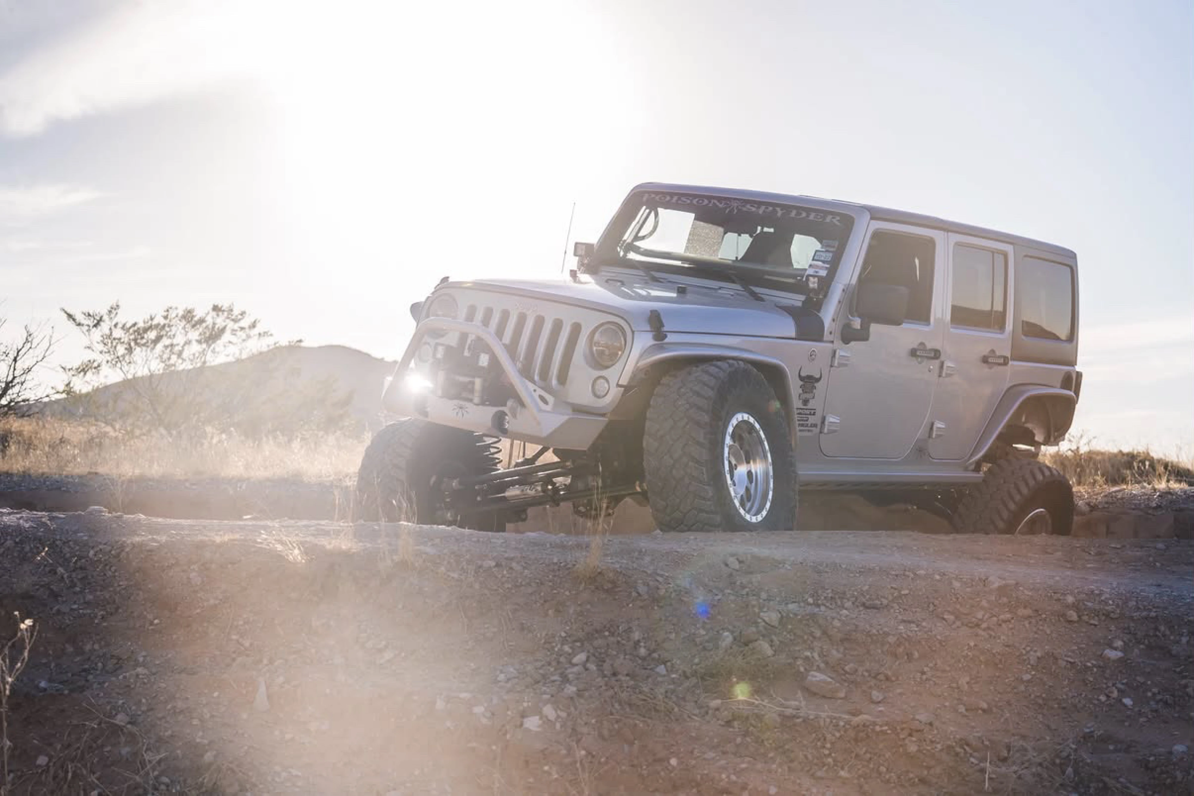 Jeep Wrangler JK driving through dust at golden hour, front three-quarter view with its Poison Spyder Brawler-Lite bumper and Crusher flares, lens flare